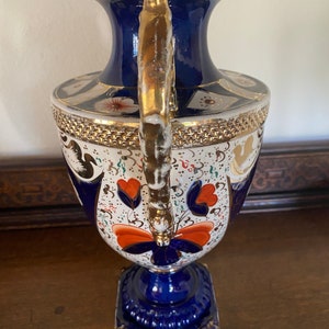 Pode incluir: Um vaso de porcelana azul e branco com detalhes dourados e um design floral. O vaso tem uma base quadrada e um corpo redondo com um pesco&ccedil;o estreito. A al&ccedil;a &eacute; dourada e tem um design decorativo.