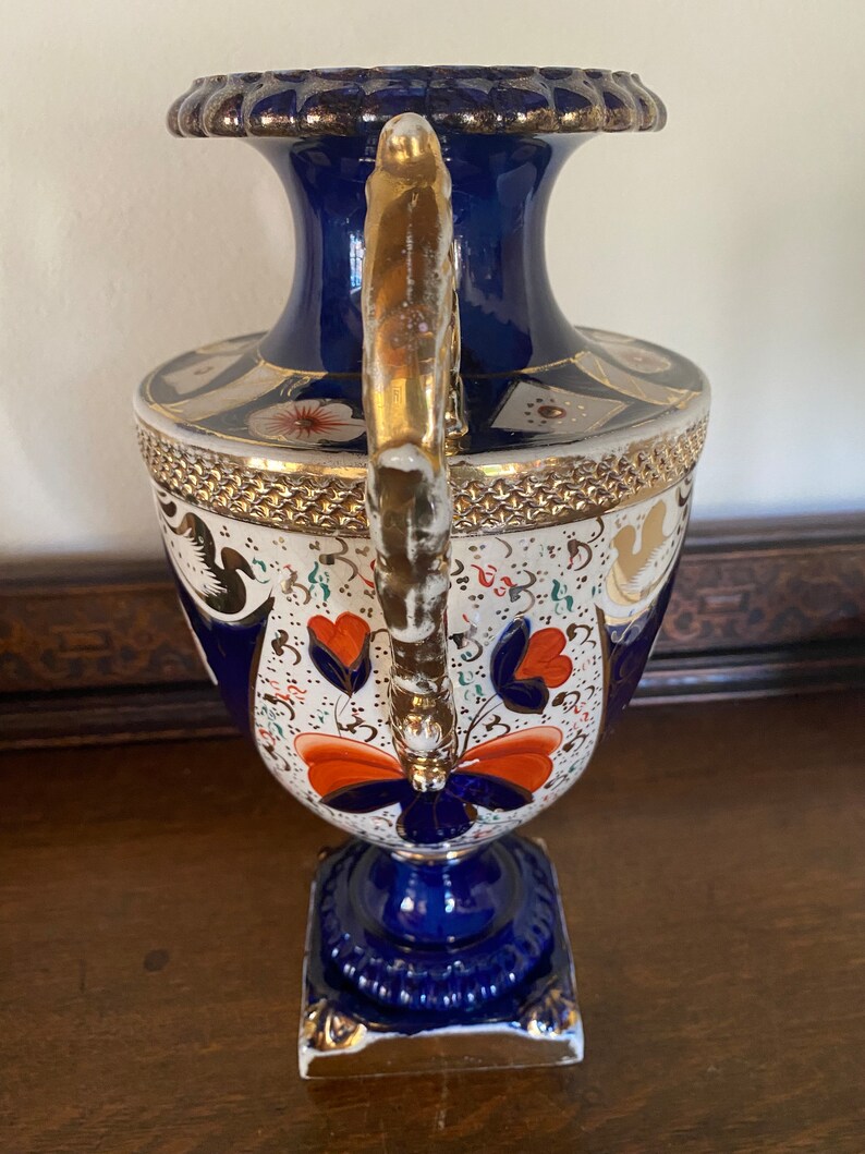 Pode incluir: Um vaso de porcelana azul e branco com detalhes dourados e um design floral. O vaso tem uma base quadrada e um corpo redondo com um pesco&ccedil;o estreito. A al&ccedil;a &eacute; dourada e tem um design decorativo.