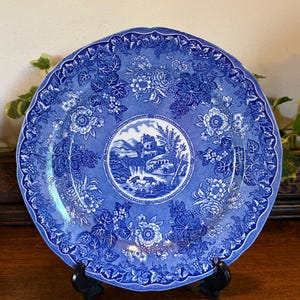 Puede incluir: Un plato decorativo azul y blanco con un diseño floral y paisajístico. El plato presenta una escena central de paisaje rodeada por un borde floral. El plato se exhibe sobre un soporte negro, mostrando sus intrincados detalles y su estética vintage.