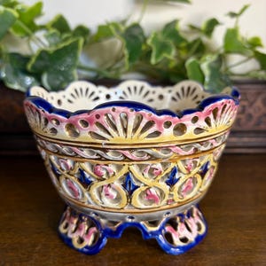 Puede incluir: Un cuenco decorativo de cerámica con borde festoneado y tres patas. El cuenco presenta un interior color crema, una banda rosa y amarilla y detalles azules. El exterior tiene un diseño calado.