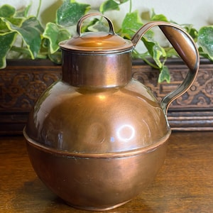 May include: A copper jug with a rounded body, a lid, and a curved handle. The jug has a shiny, reflective surface and a small handle on the lid. The copper has a warm, reddish-brown hue, and the jug appears to be vintage.