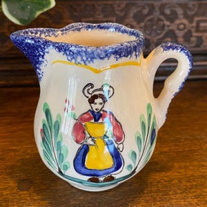 Può includere: Piccola lattiera in ceramica dipinta a mano con bordo e manico blu. La lattiera presenta un disegno di arte popolare di una donna con grembiule giallo e vestito blu, circondata da fogliame verde e rosso. La lattiera è alta circa 7,6 cm.