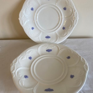May include: Two white ceramic plates with a scalloped edge and a floral design. The plates have a raised center with a smaller circle in the middle. The floral design is in shades of blue.