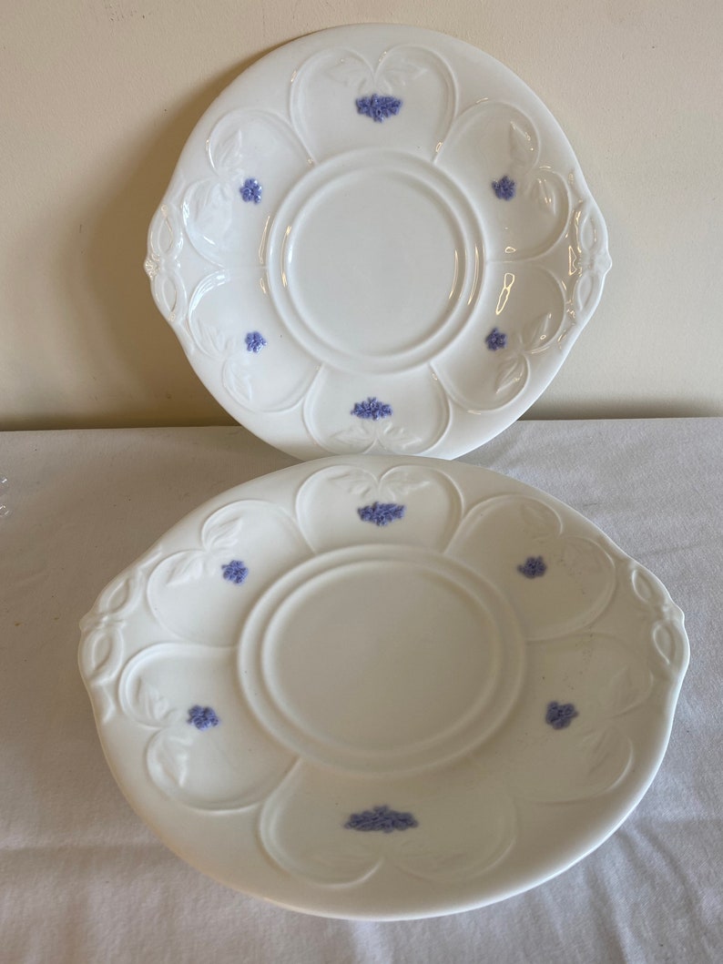 May include: Two white ceramic plates with a scalloped edge and a floral design. The plates have a raised center with a smaller circle in the middle. The floral design is in shades of blue.