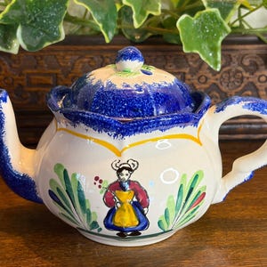 May include: A ceramic teapot with a blue and white floral design. The lid and spout are blue, and the body is white with a painted figure in traditional clothing. The handle is curved.