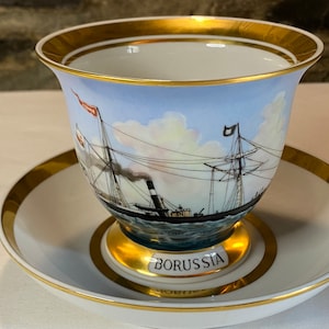 May include: A white porcelain teacup and saucer with a gold rim. The cup features a painted image of a ship with the word "BORUSSIA" on the saucer.