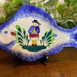 May include: A ceramic fish-shaped plate with a blue speckled border. The plate features a painted scene of a person in traditional clothing playing a drum, surrounded by green foliage. The plate is supported by a black stand.