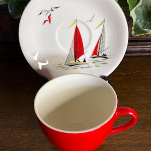 May include: A vintage teacup and saucer set. The red teacup is paired with a white saucer featuring a nautical design of sailboats with red sails and seagulls. The set has a gold trim.