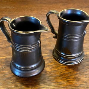 Könnte beinhalten: Zwei kleine braune Keramikkrüge mit goldener Verzierung. Die Krüge haben einen runden Körper und einen Henkel.