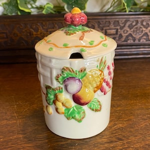 May include: A cream ceramic jar with a lid and a small spoon opening. The jar is adorned with colourful fruit and green leaves. The lid is topped with a red and yellow flower-shaped knob. The jar is likely for jams or preserves, a kitchen item.