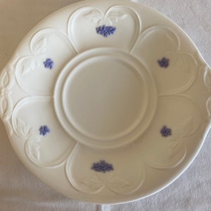 May include: A white ceramic saucer with a raised design and purple floral accents. The saucer has a center well for a cup.