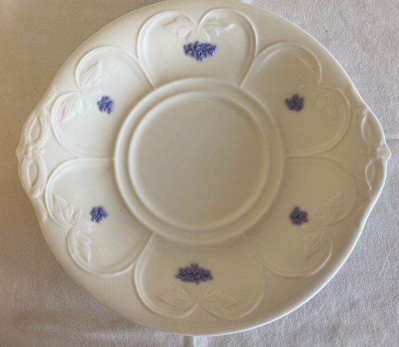 May include: A white ceramic saucer with a raised design and purple floral accents. The saucer has a center well for a cup.