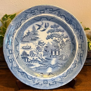 Op de afbeelding: Een ronde, blauw-witte decoratieve plaat met een gedetailleerde landschapsscène. De plaat heeft pagodes, bruggen en bomen, met een rand van ingewikkelde patronen. De plaat wordt gepresenteerd op een houten standaard.