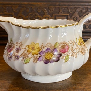 Könnte beinhalten: Eine weiße Keramik-Milchkanne mit goldfarbenem Rand und Blumenmuster. Die Kanne hat ein geriffeltes Design und einen gebogenen Griff.