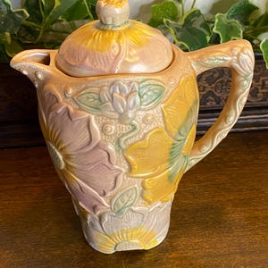 Pode incluir: Um cremeira de cerâmica com tampa, com desenhos florais em relevo em tons de amarelo, rosa e verde. A alça e o bico são integrados no design. A cremeira é uma peça decorativa para servir bebidas.