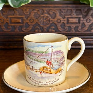 May include: A cream-coloured ceramic cup and saucer set with a gold rim. The cup features a hand-painted illustration of a chairlift in a snowy mountain landscape, with the text "THE CAIRNGORM CHAIR-LIFT NEAR AVIEMORE, INVERNESS-SHIRE" printed below.