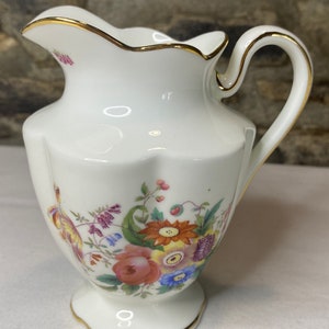 Könnte beinhalten: Eine weiße Porzellan-Milchkanne mit goldfarbenem Rand und Blumenmuster. Die Kanne hat einen gewellten Rand und einen zarten Griff.