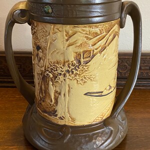 May include: A brown ceramic vase with two handles and a relief design of a scene with a man fishing in a lake. The vase has a brown glaze with a yellow glaze on the relief design.