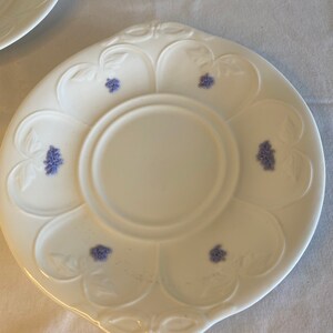 May include: A white porcelain saucer with a scalloped edge and a floral design. The saucer has a center well and is decorated with small purple flowers.