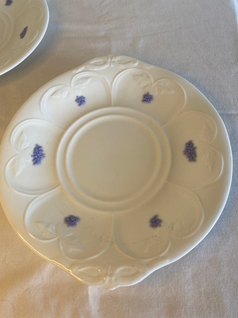 May include: A white porcelain saucer with a scalloped edge and a floral design. The saucer has a center well and is decorated with small purple flowers.