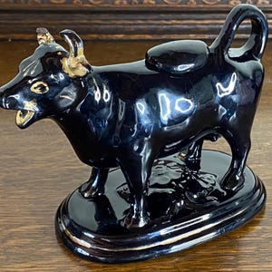May include: Black ceramic creamer in the shape of a bull with gold accents. The creamer has a round base and a spout on the back.
