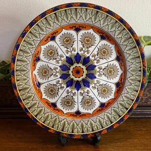 May include: A decorative plate with a floral design. The plate features a central blue and orange flower surrounded by smaller flowers in brown and white. The border has a green and orange leaf pattern with a blue and gold trim.