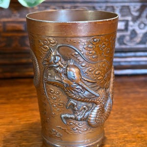 Puede incluir: Un vaso cilíndrico de metal en tono cobre que presenta un relieve detallado de un dragón que se enrosca alrededor del exterior. El dragón está representado con escamas, garras y un cuerpo largo, rodeado de motivos de nubes estilizadas.