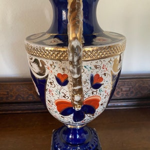 Pode incluir: Um vaso de porcelana azul e branco com detalhes dourados e desenhos florais. O vaso tem uma al&ccedil;a dourada torcida e uma base quadrada.