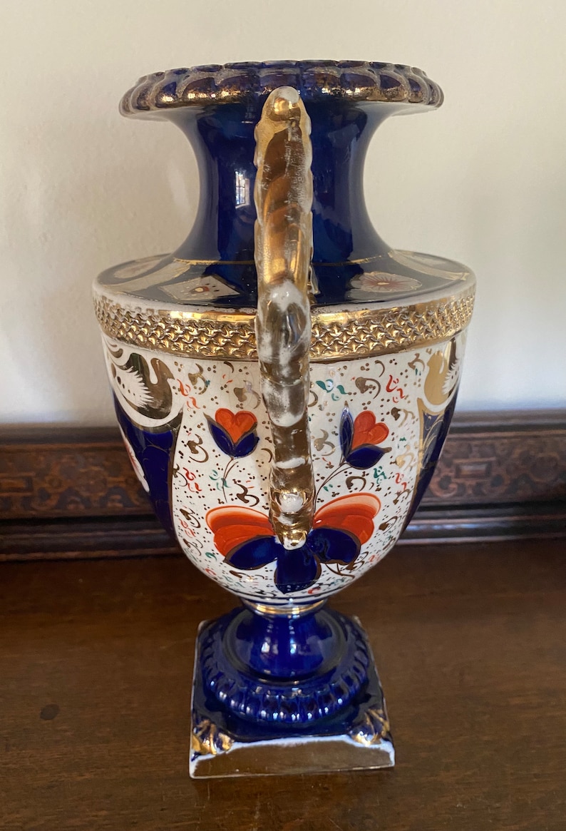 Pode incluir: Um vaso de porcelana azul e branco com detalhes dourados e desenhos florais. O vaso tem uma al&ccedil;a dourada torcida e uma base quadrada.