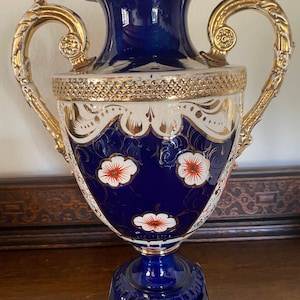 Pode incluir: Um vaso de porcelana azul e dourado com desenhos florais e detalhes dourados. O vaso tem duas al&ccedil;as e uma base quadrada.