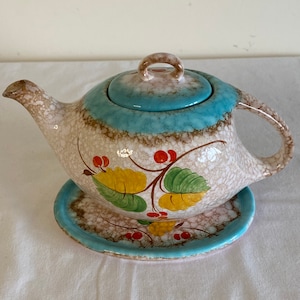 May include: A ceramic teapot with a matching saucer. The teapot has a speckled beige body with a turquoise band and lid. It is decorated with a floral design featuring yellow and green leaves and red berries. The teapot is designed for serving tea.