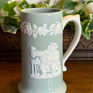 May include: A sage green ceramic tankard featuring a white relief design of a seated figure holding a pitcher at a table. The upper rim is decorated with a white embossed floral garland. The tankard has a smooth, glossy finish and a white handle.