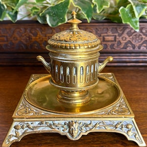 Puede incluir: Un tintero antiguo de latón con un diseño ornamentado y detallado. El tintero tiene un recipiente con tapa y asas, colocado sobre una base cuadrada con relieve decorativo. La pieza es de color dorado cálido, lo que sugiere antigüedad y artesanía.
