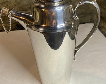 May include: A silver cocktail shaker with a handle and a spout, featuring a lid and a chain. The shaker has a polished finish and a classic design, suitable for mixing drinks. The shaker is approximately 10 inches tall.