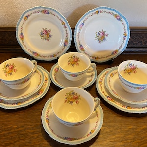 Può includere: Un set di sei tazze da tè e piattini vintage con motivi floreali. Le tazze e i piattini sono bianchi con bordi blu e dorati. I motivi floreali sono in tonalità di rosa, giallo e verde.