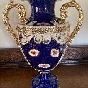 Pode incluir: Um vaso de porcelana azul e branco com detalhes dourados e desenhos florais. O vaso tem duas al&ccedil;as e uma base quadrada.