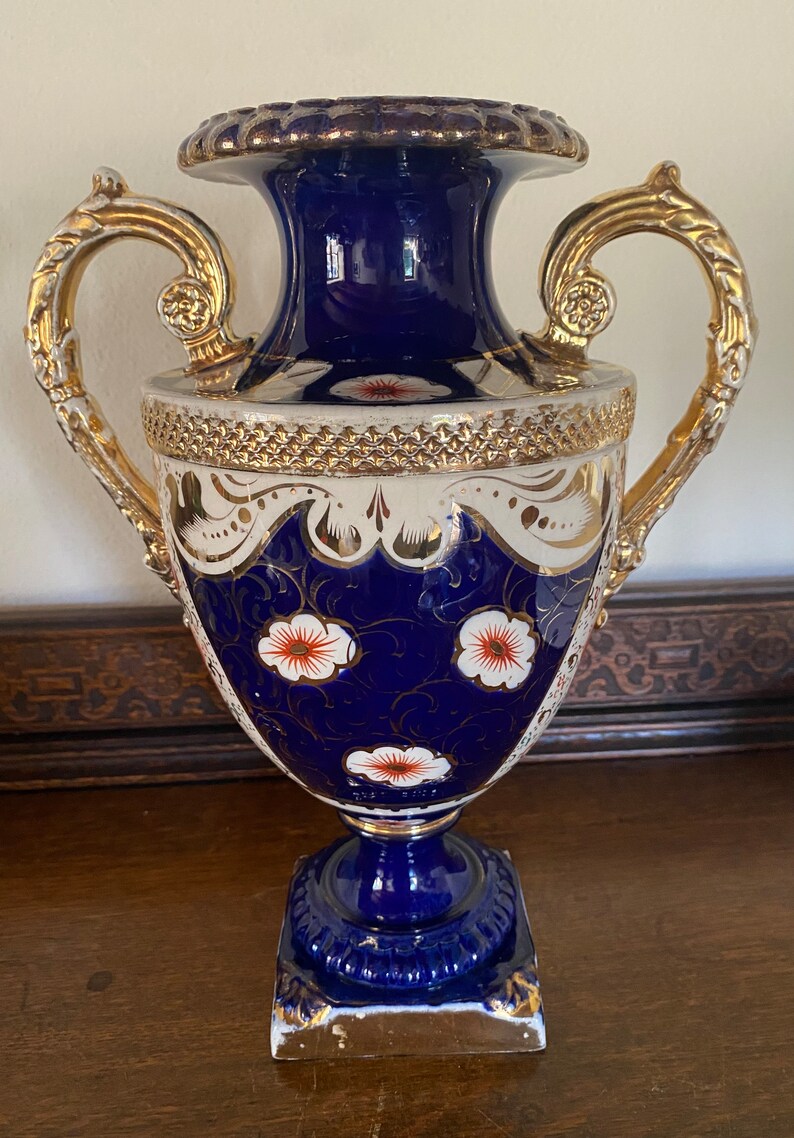 Pode incluir: Um vaso de porcelana azul e branco com detalhes dourados e desenhos florais. O vaso tem duas al&ccedil;as e uma base quadrada.