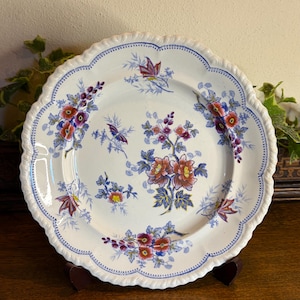 Puede incluir: Un plato de cerámica blanca con borde festoneado y borde azul punteado. El plato presenta un diseño floral con flores y hojas rojas, moradas y azules. El plato se exhibe sobre un soporte de madera.