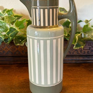 May include: A ceramic jug with a green and white striped pattern. The jug features a rounded body, a spout, and a curved handle. The color palette is predominantly green and white, with a darker green accent on the top and base.