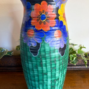 May include: A ceramic vase with a flared rim, featuring a black upper section decorated with vibrant orange, blue, and yellow flowers. The lower portion of the vase has a textured, grid-like pattern in shades of green, creating a distinct contrast.