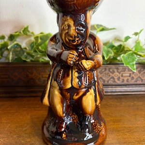 Puede incluir: Figura de cerámica marrón de un hombre con sombrero y abrigo. La figura tiene un acabado brillante con tonos marrones y beige. El rostro del hombre muestra una sonrisa, con las manos juntas. Es un objeto decorativo, de unos 15 cm de altura.