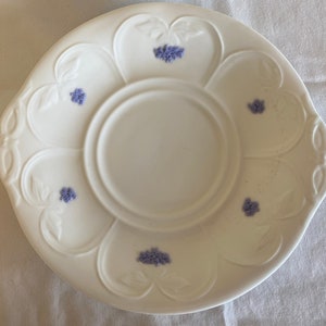 May include: White ceramic saucer with a scalloped edge and a central well. The saucer is decorated with small purple floral designs.
