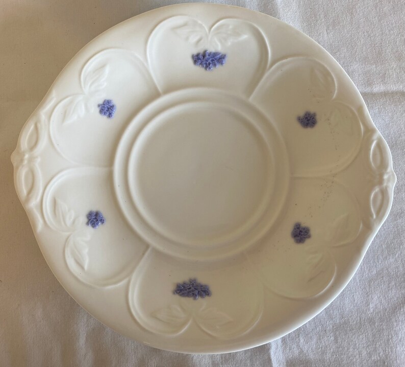 May include: White ceramic saucer with a scalloped edge and a central well. The saucer is decorated with small purple floral designs.