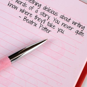 May include: A pink pen rests on a lined pink notebook with the quote "Something delicious about writing words of a story. You never quite know where they'll take you. - Beatrix Potter" written in black ink.