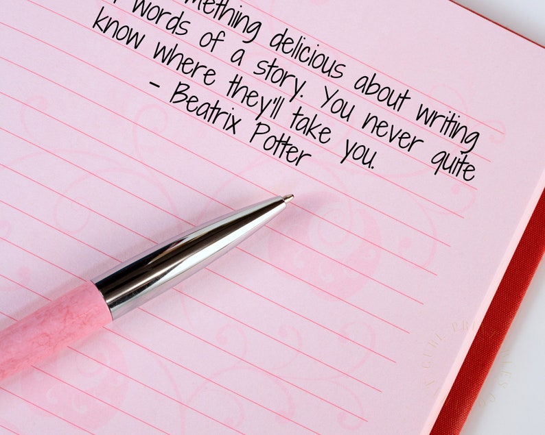 May include: A pink pen rests on a lined pink notebook with the quote "Something delicious about writing words of a story. You never quite know where they'll take you. - Beatrix Potter" written in black ink.