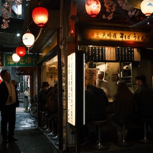 Puede incluir: Un callej&oacute;n poco iluminado en Jap&oacute;n con linternas rojas y blancas colgando sobre un restaurante. El restaurante tiene un letrero que dice "Izakaya", que es un tipo de pub japon&eacute;s. Hay gente sentada en mesas dentro del restaurante.