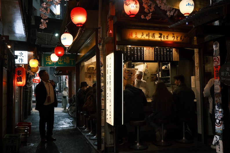 Puede incluir: Un callej&oacute;n poco iluminado en Jap&oacute;n con linternas rojas y blancas colgando sobre un restaurante. El restaurante tiene un letrero que dice "Izakaya", que es un tipo de pub japon&eacute;s. Hay gente sentada en mesas dentro del restaurante.