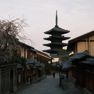 Puede incluir: Una pagoda japonesa tradicional con un acabado de madera oscura se alza sobre un cielo nublado. La pagoda est&aacute; rodeada de una calle estrecha bordeada de edificios tradicionales japoneses.