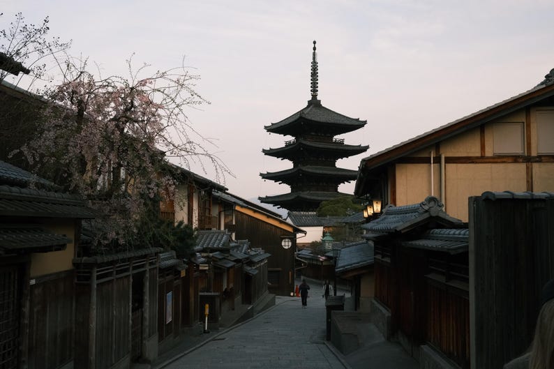 Puede incluir: Una pagoda japonesa tradicional con un acabado de madera oscura se alza sobre un cielo nublado. La pagoda est&aacute; rodeada de una calle estrecha bordeada de edificios tradicionales japoneses.