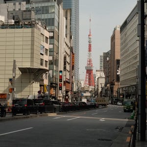 Puede incluir: Una vista de una calle de la ciudad de Tokio, Jap&oacute;n, con una alta torre roja y blanca en la distancia. La torre es visible entre los edificios y es el punto focal de la imagen. La calle est&aacute; bordeada de edificios y coches.